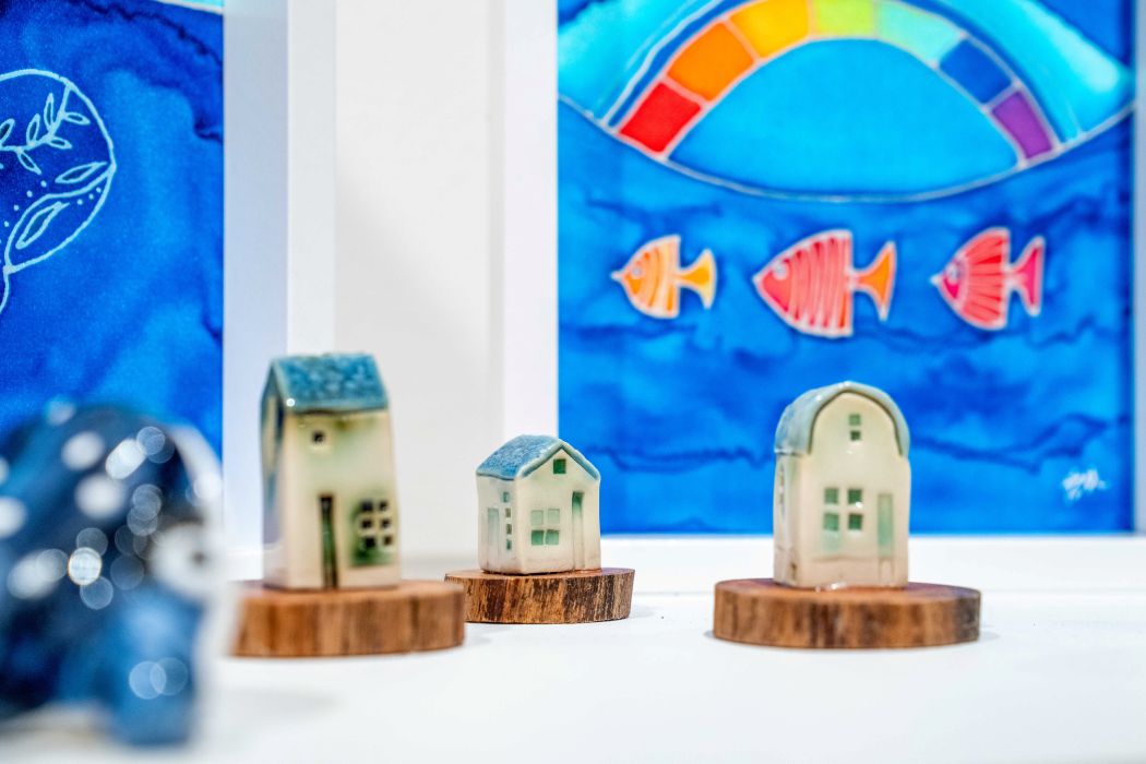 Three small ceramic houses on wooden blocks sit in the foreground, with blue artwork behind them showing red and orange fish